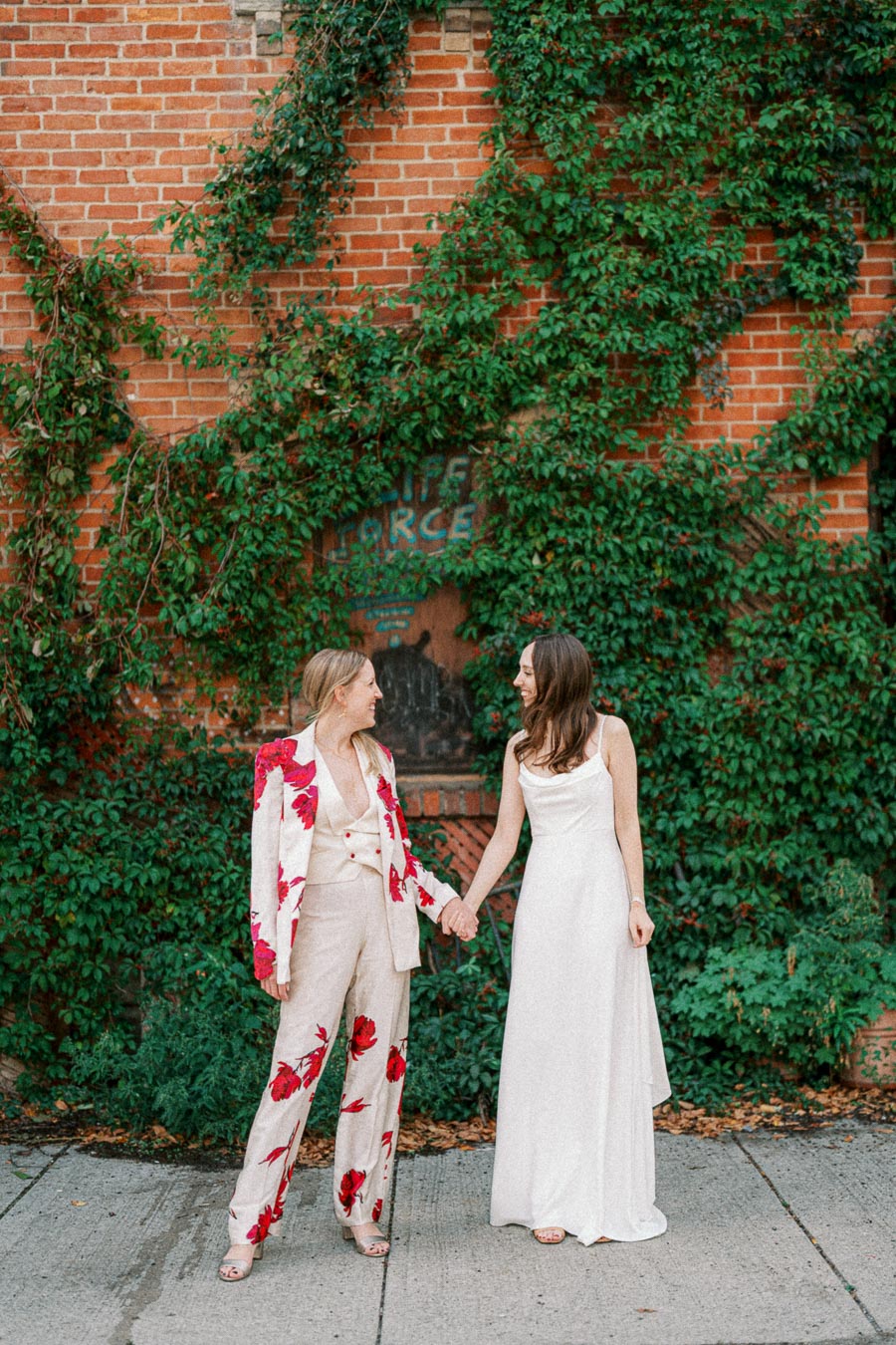 Two individuals standing hand in hand against a rustic brick wall covered in green ivy, one wearing a floral suit and the other in a white dress, embodying style and elegance in an outdoor setting.