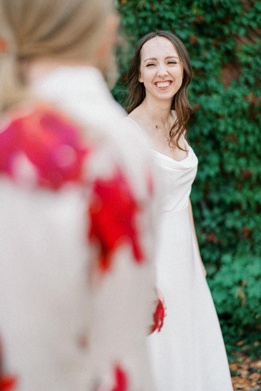 Smiling bride in white dress looking at partner with blurred floral pattern in the foreground, standing against a lush green leafy background.