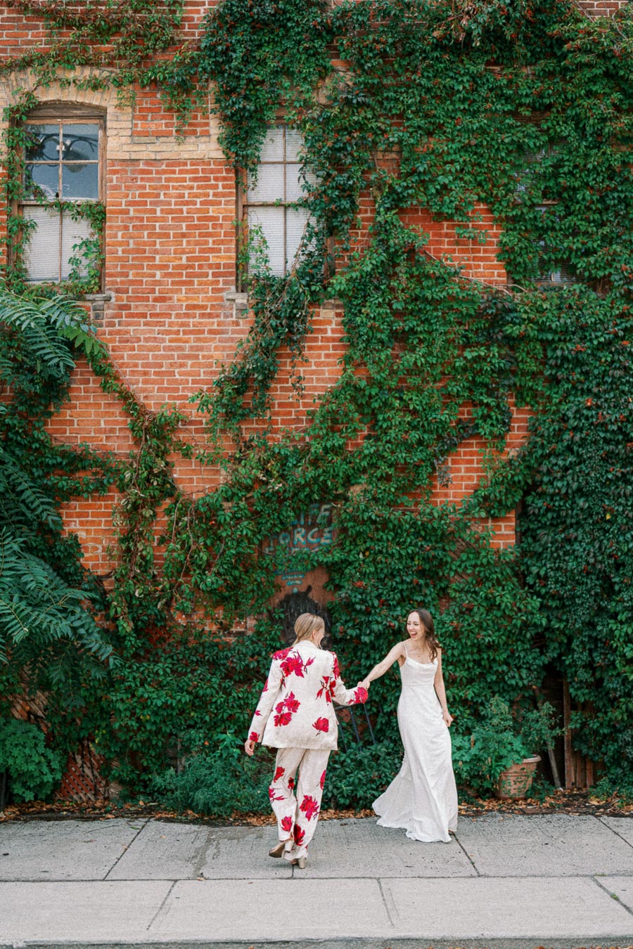 Couple joyfully dancing in front of a brick wall covered in lush green ivy, with one person wearing a floral suit and the other in a white dress.
