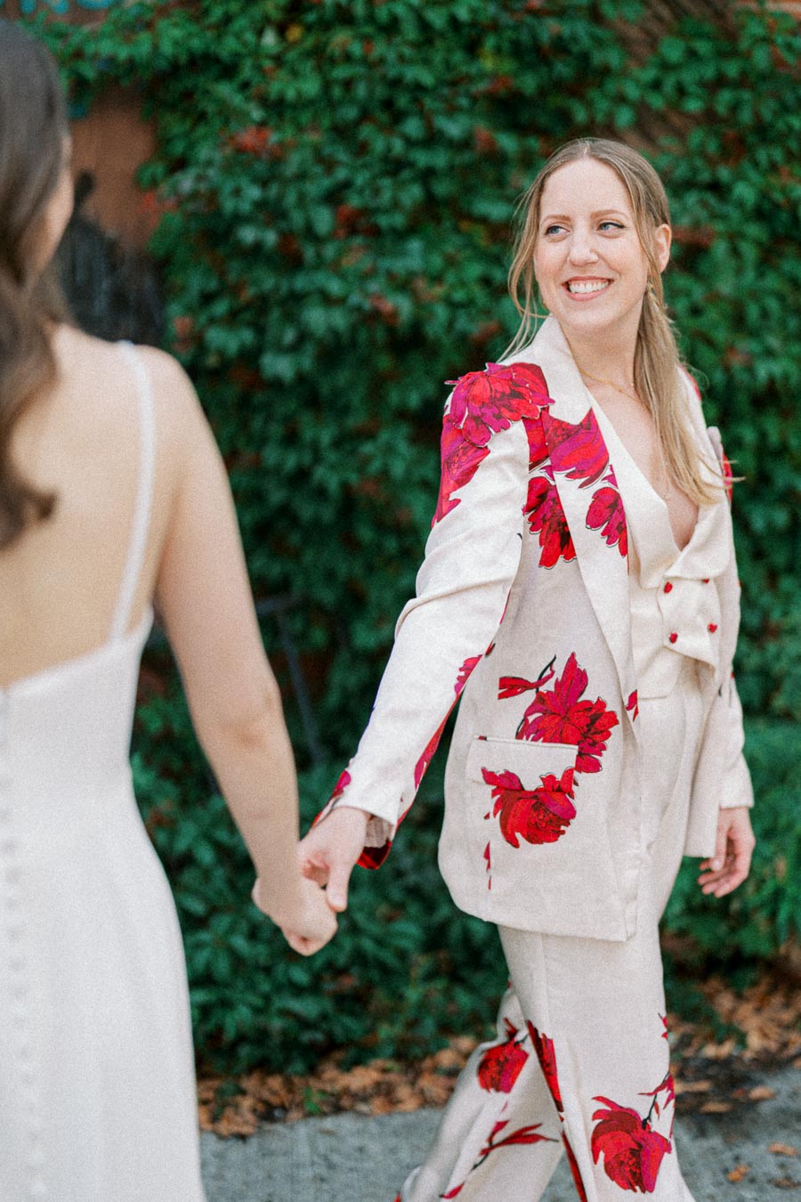 A woman in a stylish suit with red floral patterns holds hands with another woman in a white dress, walking outdoors in front of lush greenery.
