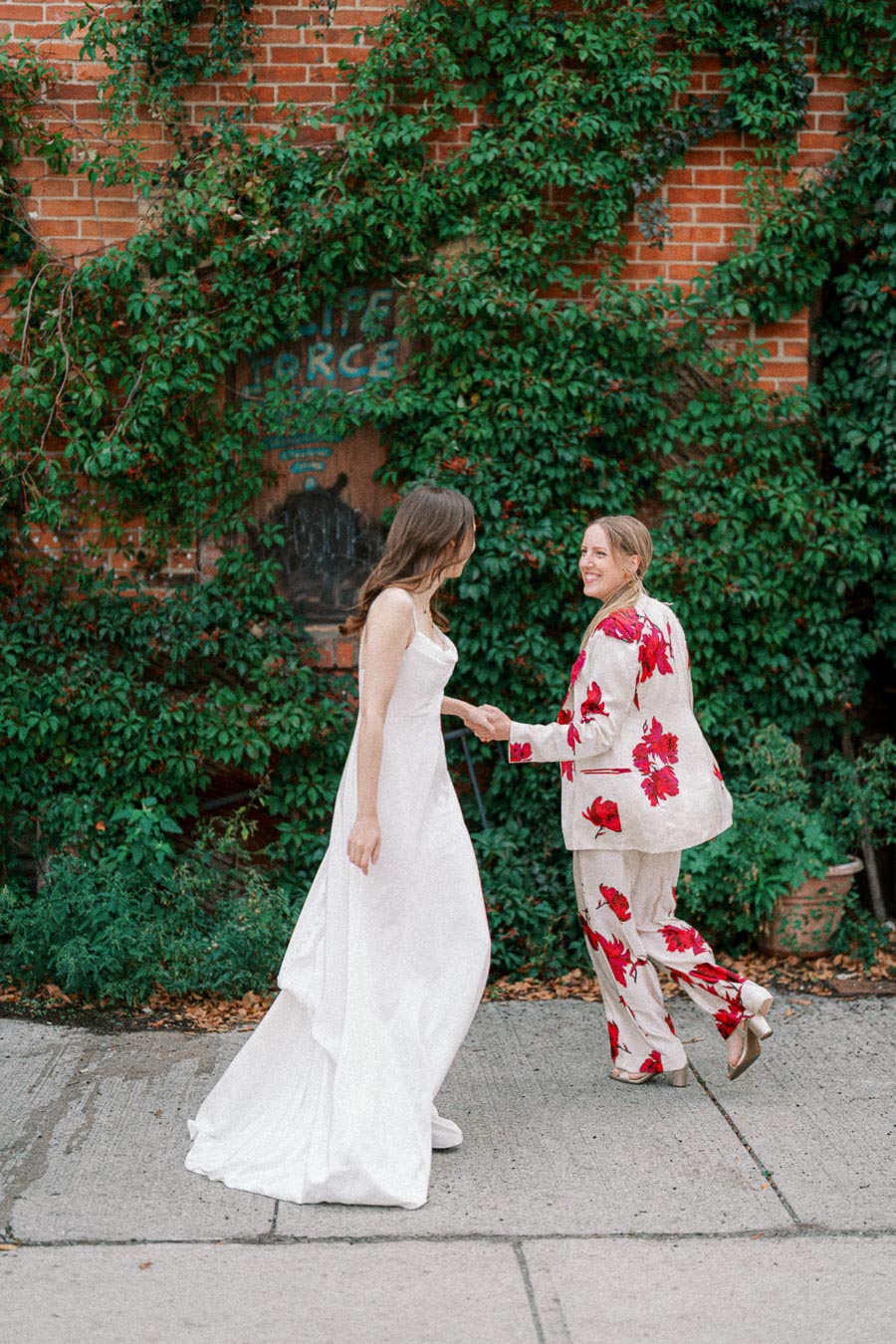 Two women joyfully holding hands in front of a lush ivy-covered brick wall; one wearing a white wedding dress and sneakers, the other in a floral suit, creating a vibrant and playful scene.