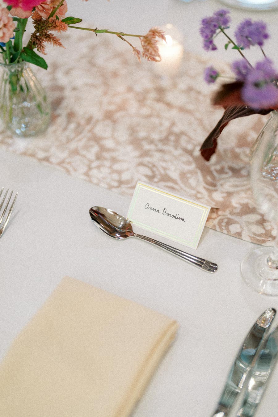 Elegant wedding table setting featuring a floral centerpiece, a place card, and polished cutlery on a lace tablecloth.