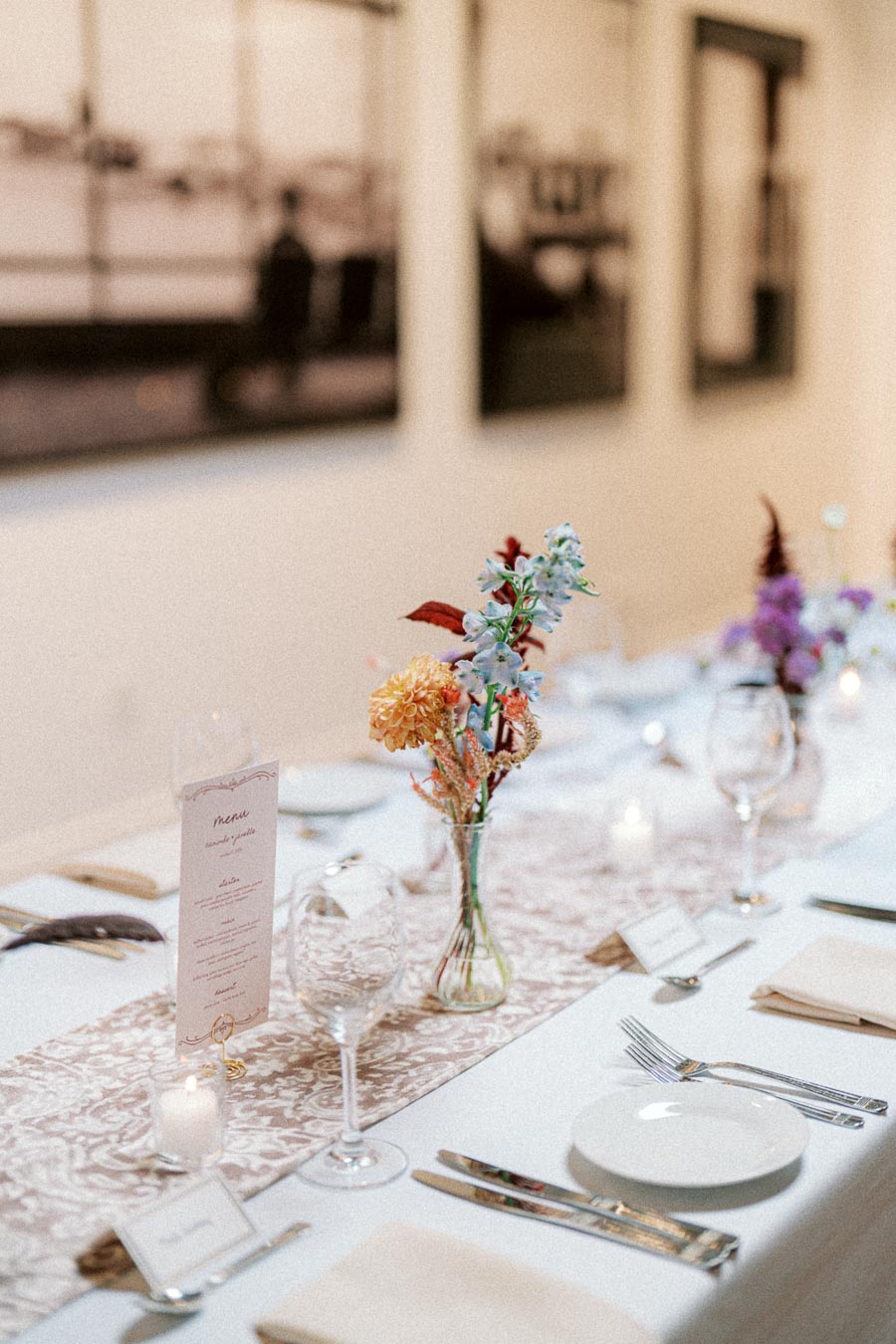 Elegant dining table setting with floral centerpiece, wine glasses, plates, and cutlery, featuring soft lighting and artistic wall decor in the background.