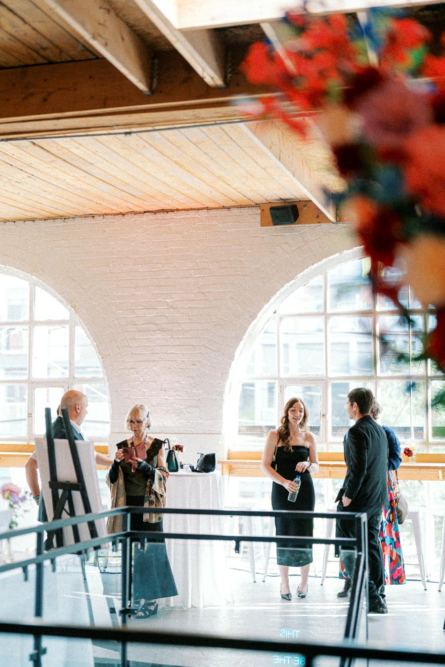 A group of people interacting at an indoor event venue with large arched windows and a high ceiling, featuring vibrant floral arrangements in the foreground.