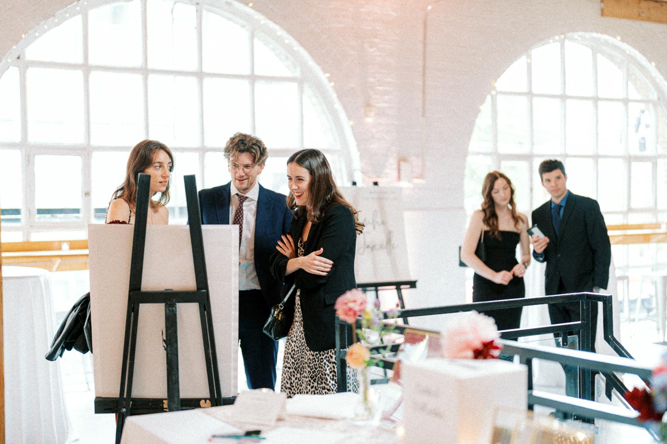 A group of people in formal attire gather around an easel display at a bright, elegant event venue with large arched windows.