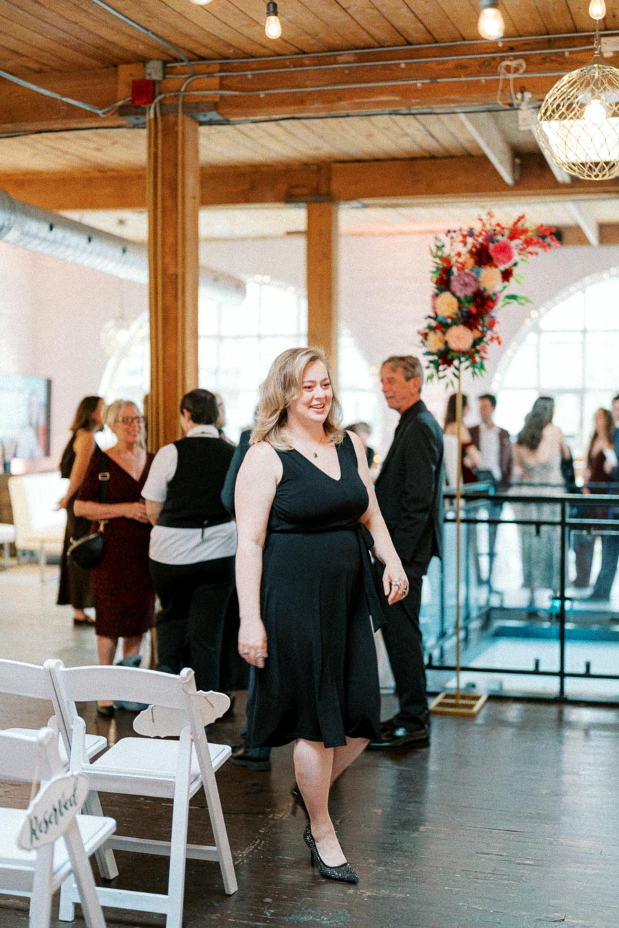 Elegant woman in a black dress smiling at a formal event, with guests mingling in the background and a vibrant floral arrangement nearby.