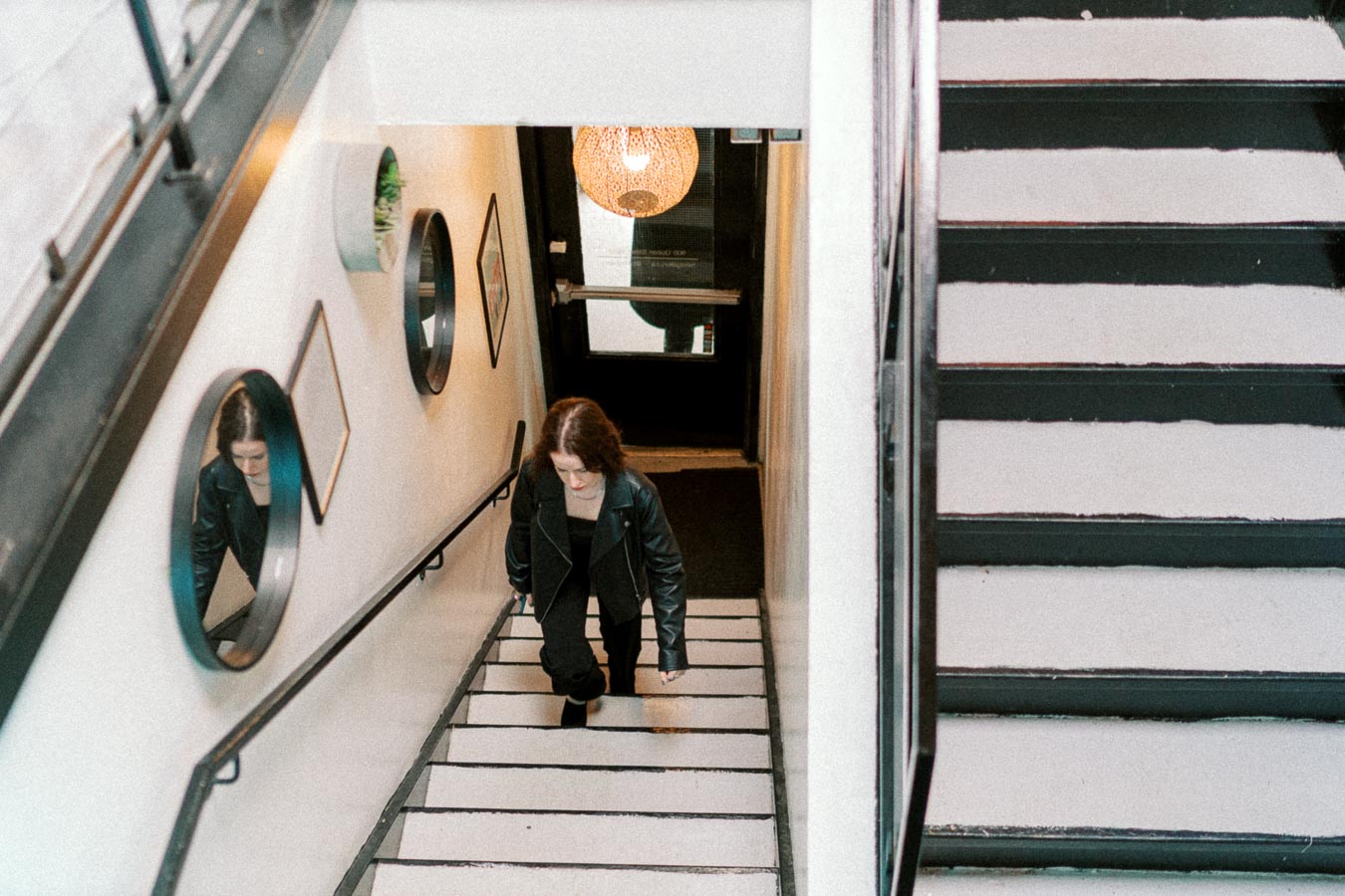 A person in a black leather jacket ascending a modern staircase with a white wall, featuring circular mirrors and frames, under soft ambient lighting.