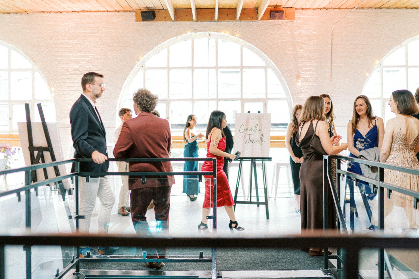 Group of elegantly dressed people socializing at a stylish indoor event with large arched windows in the background, a sign with calligraphy, and a modern, sophisticated ambience.