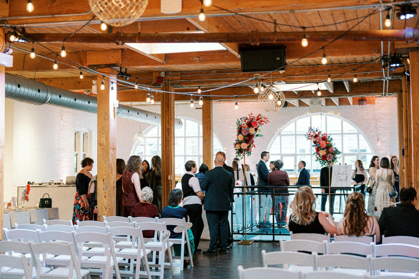 Elegant indoor wedding venue with guests mingling in a beautifully decorated space, featuring wooden beams, string lights, and vibrant floral arrangements.