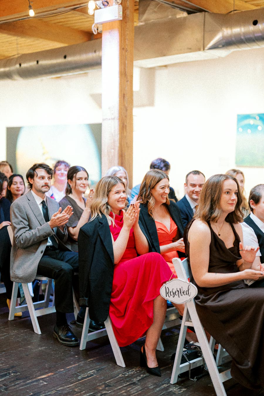 Audience clapping and smiling at an indoor event, seated in a brightly lit room with wooden flooring and contemporary decor.