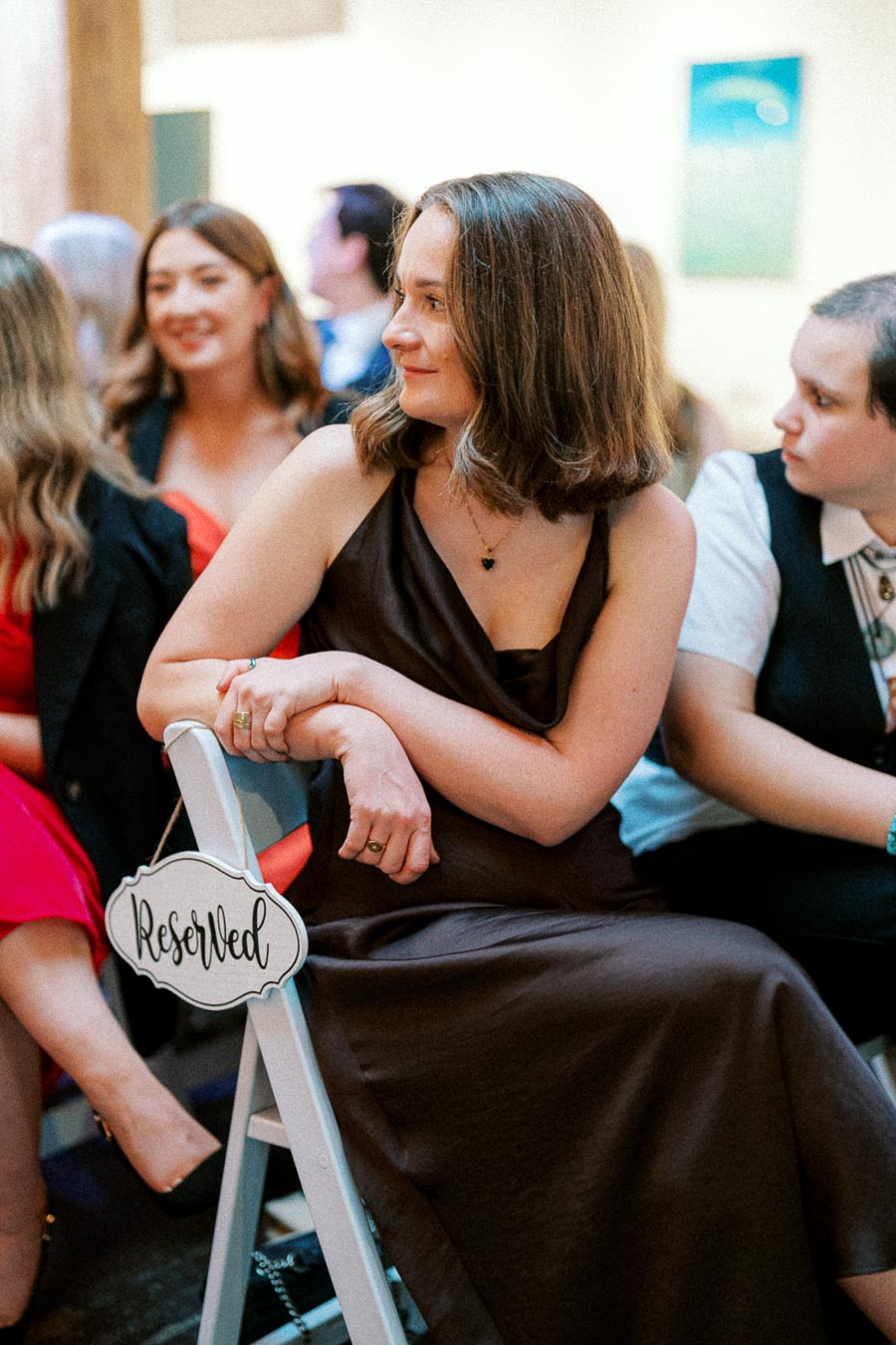 Woman seated on a white folding chair with a Reserved sign, wearing a dark sleeveless dress, at an indoor event with blurred attendees in the background.
