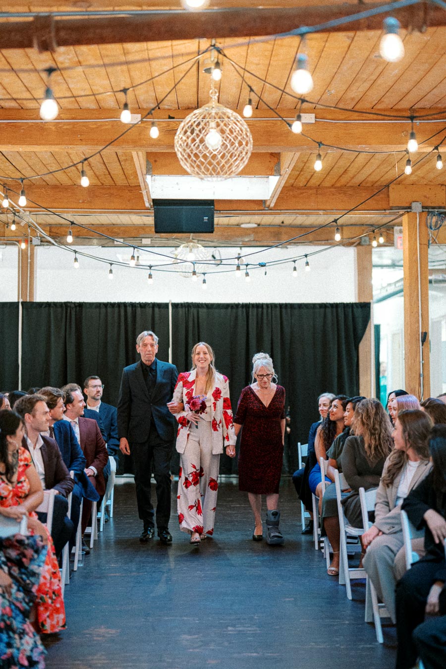 A stylish couple walks down the aisle at a wedding ceremony, set in a rustic venue adorned with string lights, surrounded by smiling guests seated on each side.