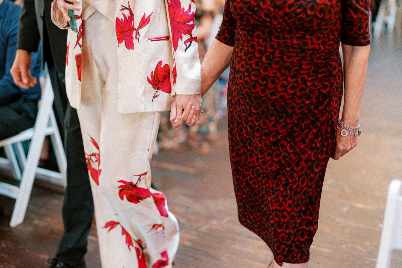 A couple holding hands while walking, dressed in stylish red and white floral and patterned attire, capturing an elegant moment at a formal event.