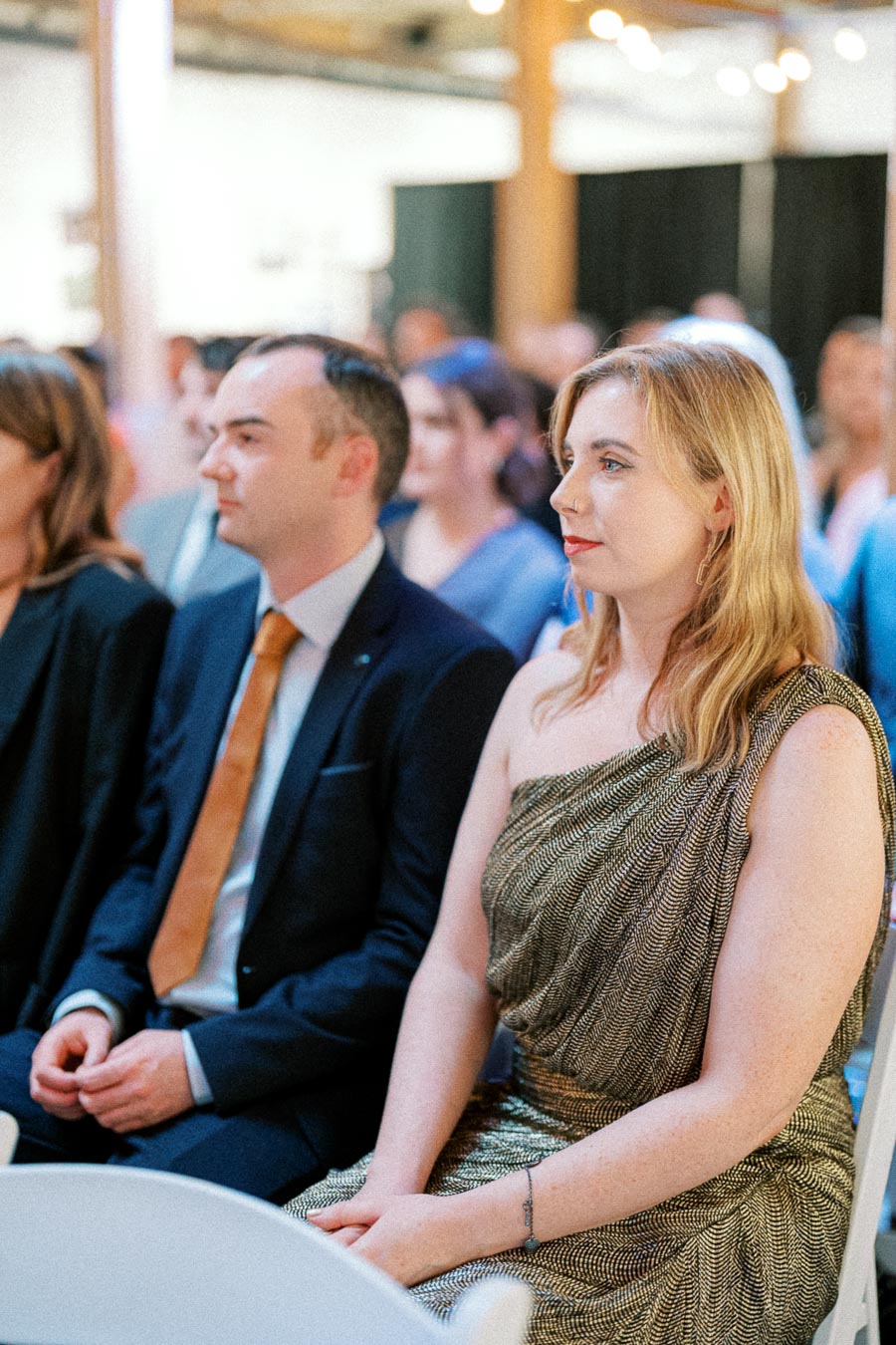 Audience watching a formal event, with a woman in a gold dress and a man in a suit seated attentively in a well-lit venue setting.