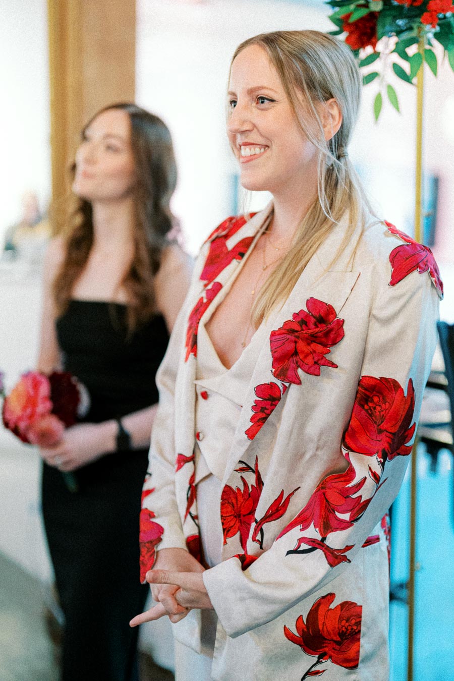 Smiling woman in a floral suit, standing beside another woman holding flowers, during a formal event.