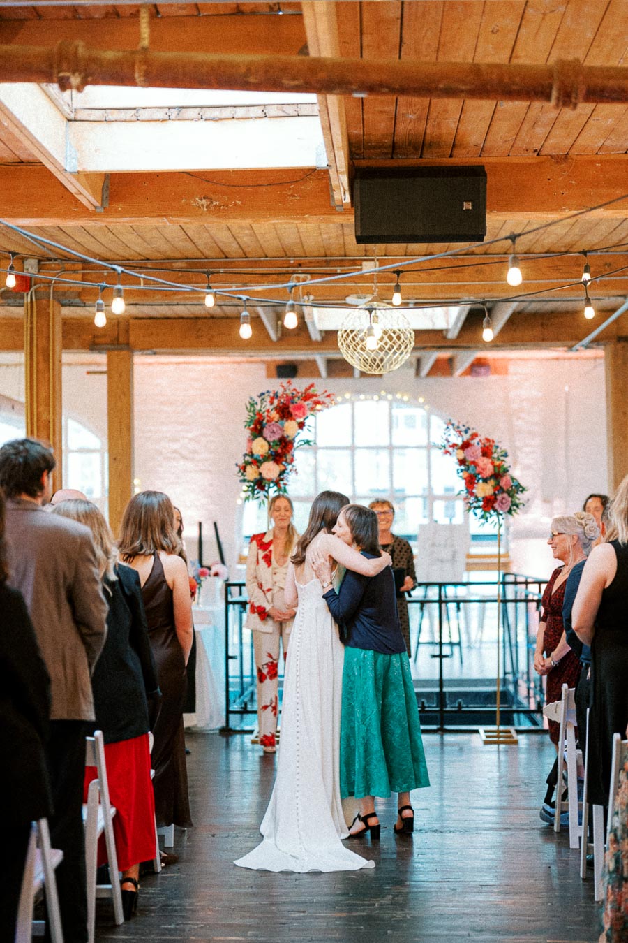 Wedding ceremony inside a rustic venue, featuring a bride in a white dress hugging a guest. The room has wooden beams, string lights, and floral arrangements, with guests in formal attire watching the embrace.