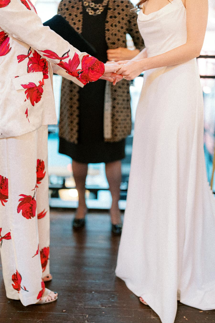 A couple holding hands during a wedding ceremony, with one wearing a floral suit and the other in a white dress, standing in front of an officiant.