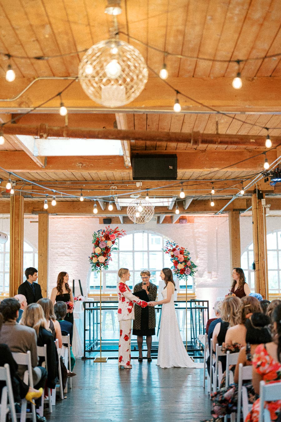 A vibrant wedding ceremony in a stylish loft venue, featuring a couple exchanging vows in front of a floral arch with colorful blossoms. An officiant is present between the couple, while guests are seated on white chairs, surrounded by rustic wood decor and hanging string lights.