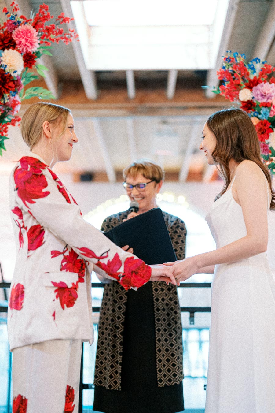 A wedding ceremony with two brides exchanging vows, one in a floral suit and the other in a white dress, while an officiant conducts the ceremony in a beautifully decorated venue with vibrant floral arrangements.