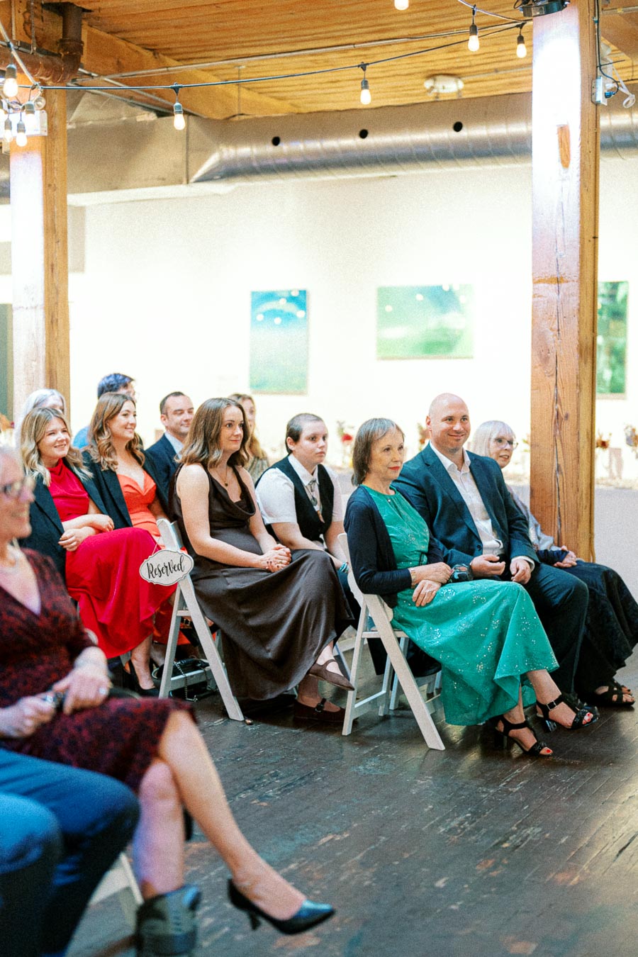A group of elegantly dressed people seated on white chairs at an indoor event, with a sign reading Reserved visible on one of the chairs. The setting appears to be a modern venue with wooden floors and decorated walls.