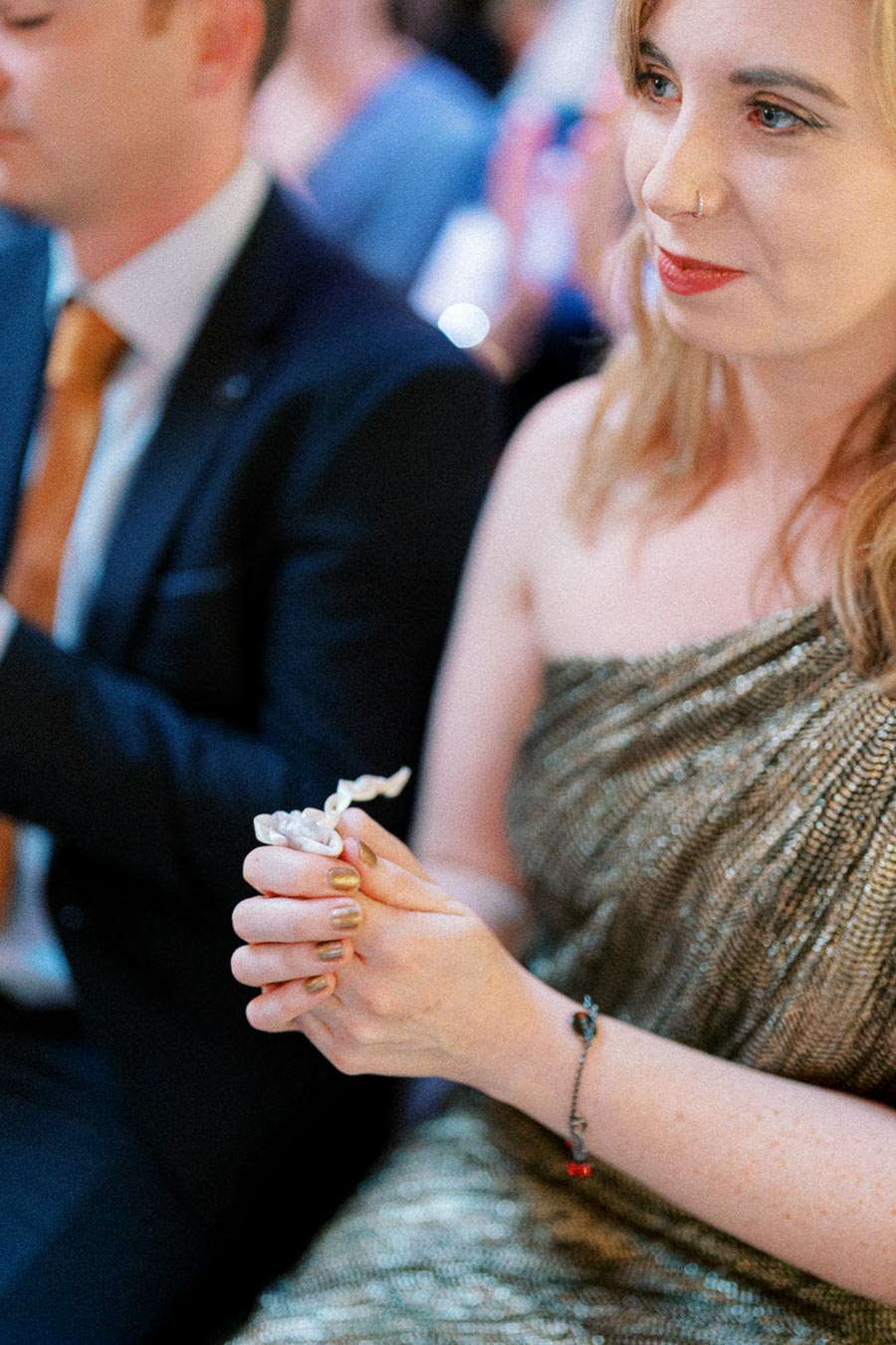 A woman in a shimmering gold dress sits attentively at an event, holding a small object in her hands.