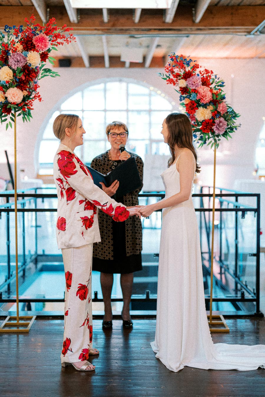A same-sex wedding ceremony with two brides holding hands, one in a floral suit and the other in a white dress. A celebrant stands behind, speaking into a microphone. The setting is modern, featuring colorful floral arrangements and a bright, spacious venue.