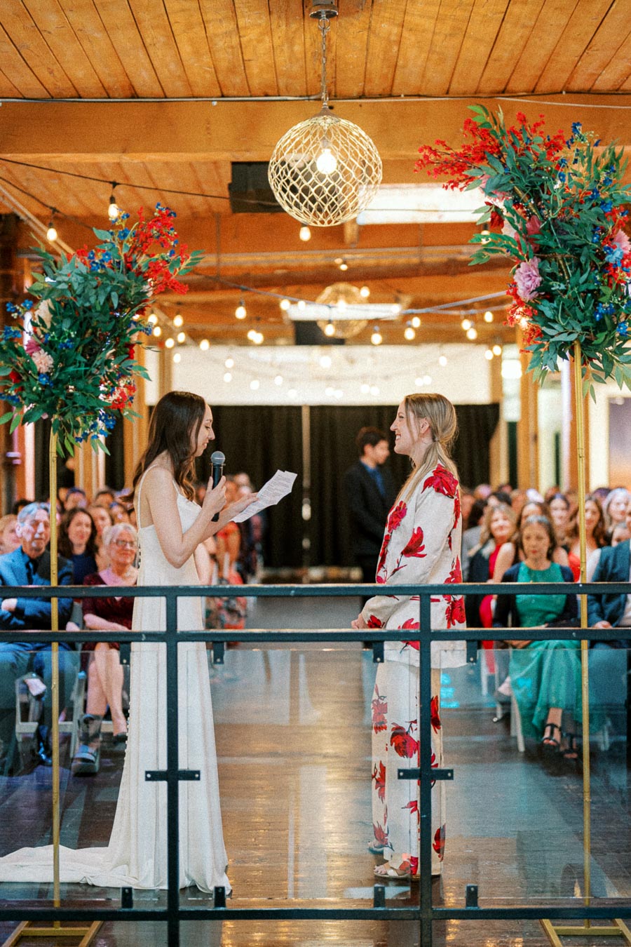 A joyful wedding ceremony with a woman in a white dress reading vows to her partner in a floral suit, surrounded by family and friends in an elegantly decorated venue.