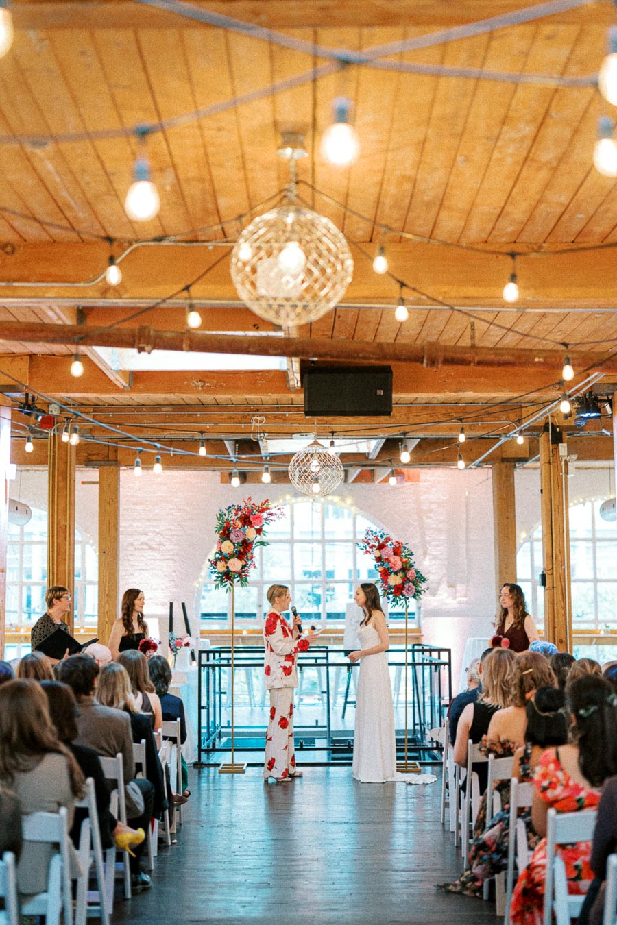 A vibrant wedding ceremony in a rustic venue, featuring two people exchanging vows under hanging string lights, surrounded by guests seated in white chairs and colorful floral arrangements.