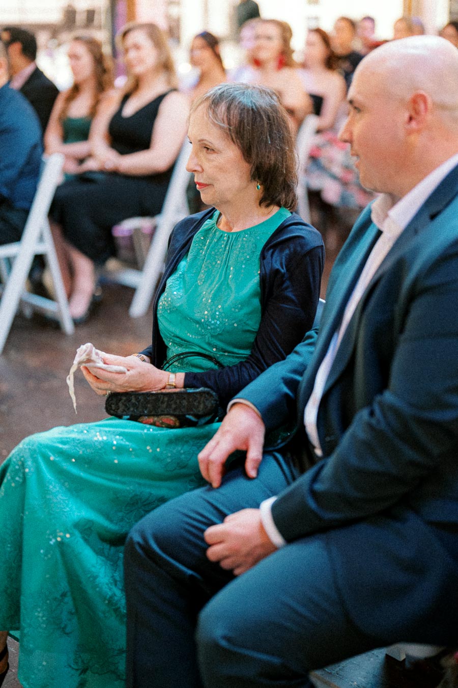 A woman in a teal dress and a man in a suit sitting attentively at an indoor event with other attendees in the background.