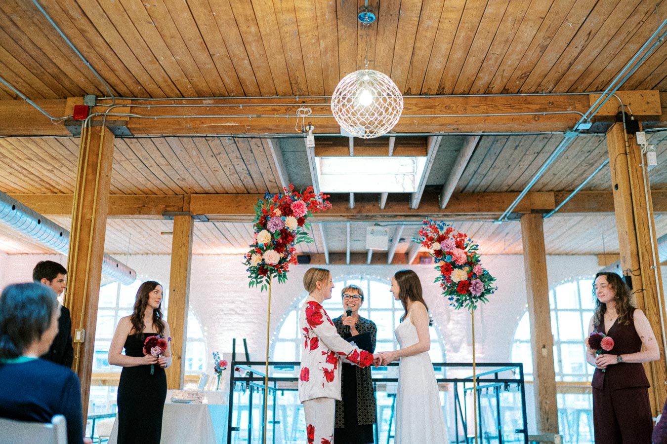 A wedding ceremony in an industrial-style venue with wooden beams and large windows. Two people stand at the altar holding hands, adorned with floral arrangements featuring vibrant red and pink flowers. An officiant is speaking to them, and guests are seated nearby.
