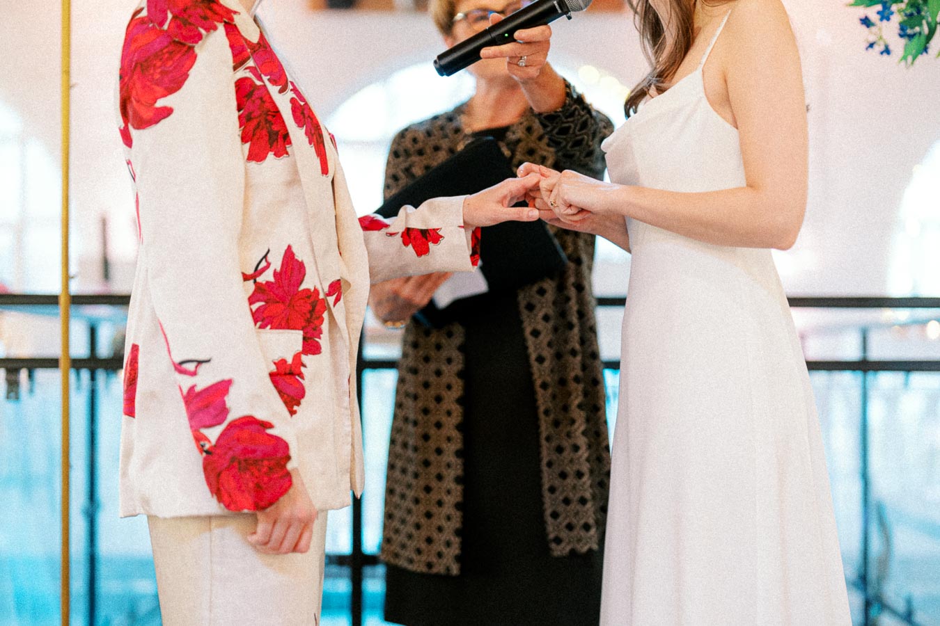 Wedding ceremony with couple exchanging rings, one wearing a floral-patterned suit, the other in an elegant white dress, while an officiant holds a microphone in the background.