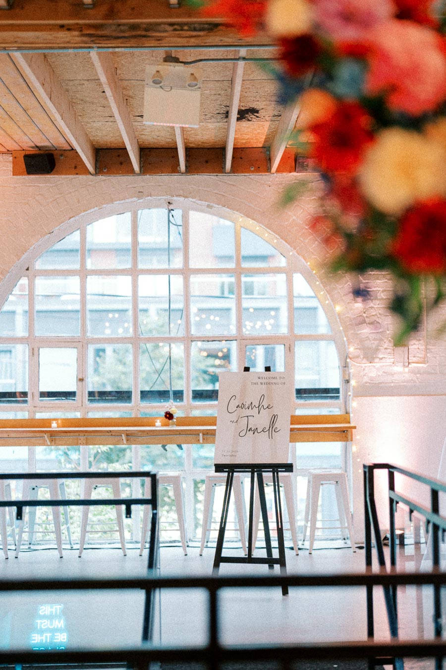 Elegant wedding venue setup featuring a welcome sign on an easel, large arched window background, flowers in the foreground, and seating arrangements at a modern urban loft.