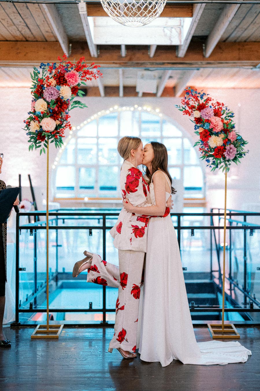 A romantic same-sex wedding ceremony with two brides kissing under a floral arch, one in a floral suit and the other in a white gown, in a bright, industrial venue.