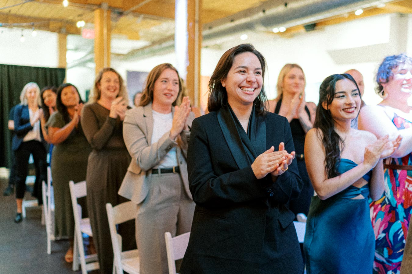 A group of diverse people standing and applauding during an indoor event, featuring individuals in formal and semi-formal attire, with a focus on one individual in a black suit smiling warmly at the front.