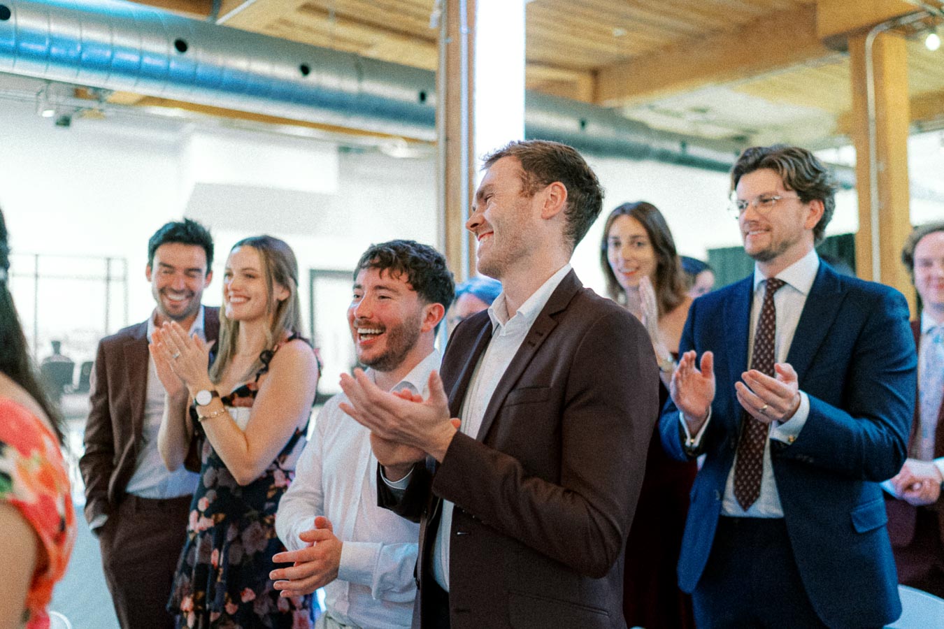 A diverse group of professionals clapping and smiling at a business event in an office setting, showcasing teamwork and positive engagement.