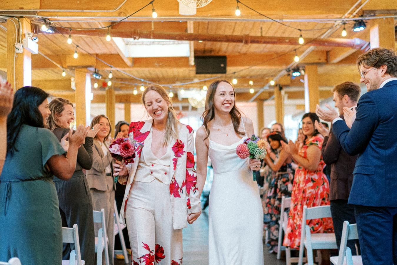 A joyful couple walking hand in hand down the aisle, surrounded by applauding guests during their wedding ceremony in a beautifully decorated venue with string lights.