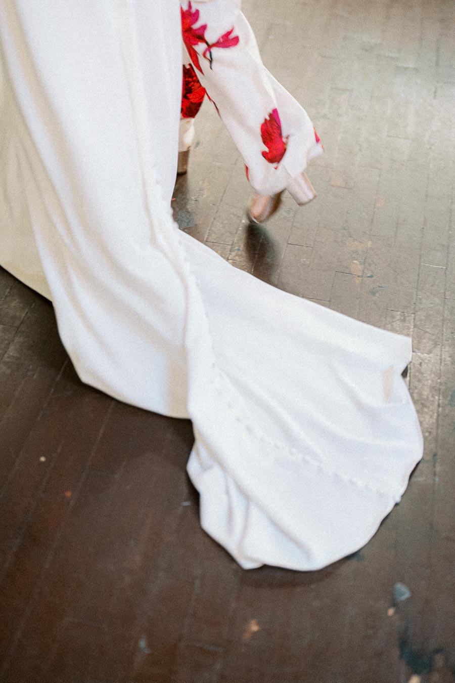 Close-up of a bride's dress train trailing elegantly on a wooden floor, with the hem of a floral patterned garment and high heels visible, capturing a stylish and romantic wedding moment.