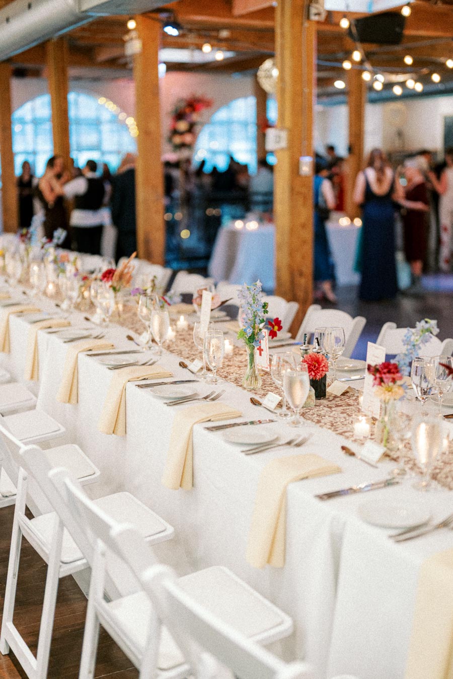 Elegant wedding reception setup with a long table adorned with white linens, floral centerpieces, candles, and neatly arranged tableware, set in a rustic venue with wooden beams and hanging string lights.
