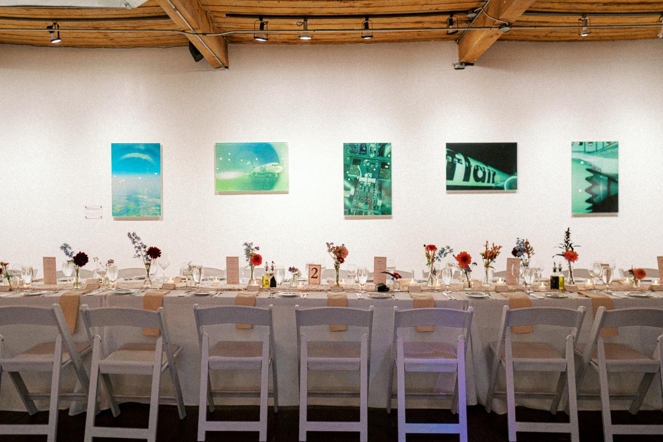 Wedding reception setup in an art gallery with a long dining table decorated with flowers and tableware, white chairs neatly arranged, and various artworks displayed on the wall.