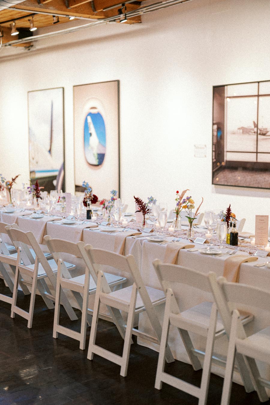 Elegant long dining table with white chairs, adorned with delicate floral centerpieces and place settings, set in a modern art gallery environment.