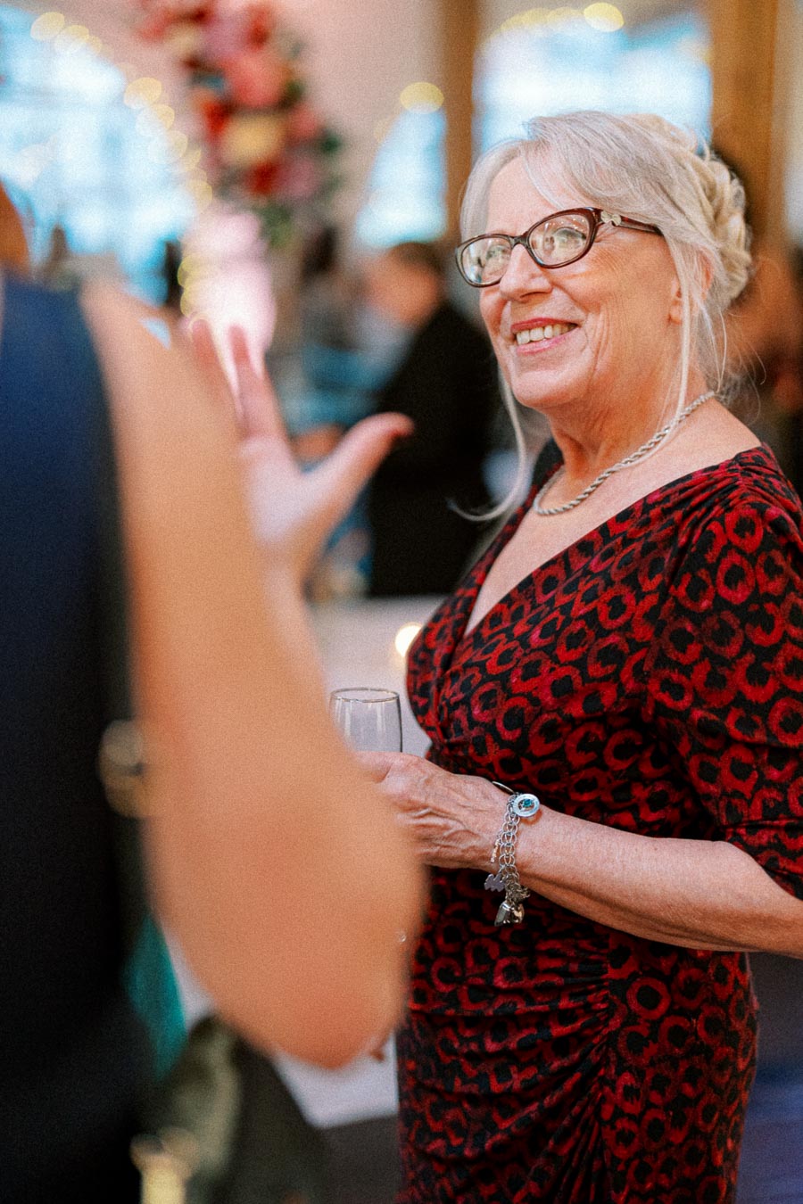 An older woman with gray hair and glasses, wearing a red and black patterned dress, smiling and holding a glass at a social event.