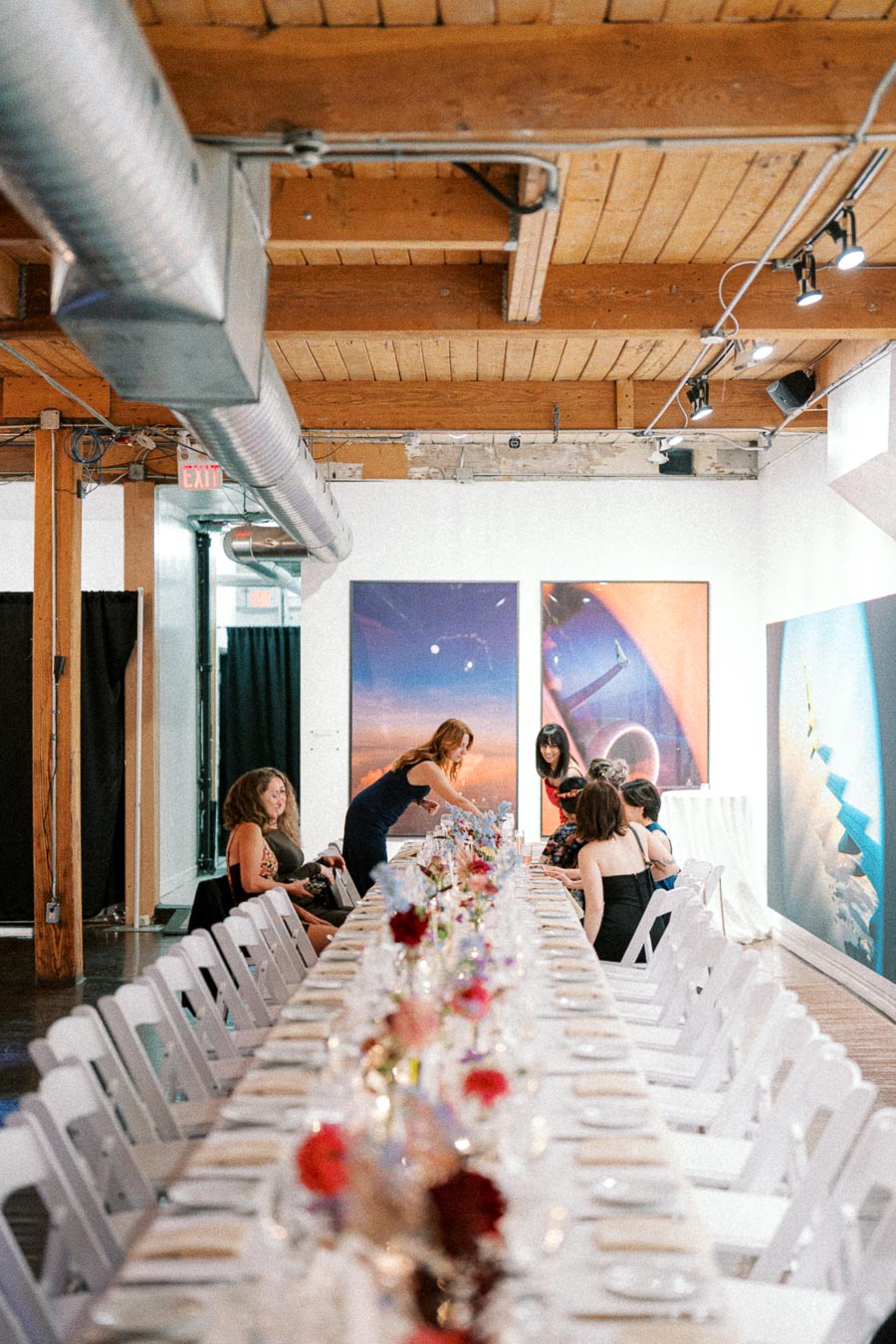Long dining table set for an elegant event in a modern loft space with wooden ceilings and industrial ductwork, featuring colorful flowers and attendees preparing for the gathering.