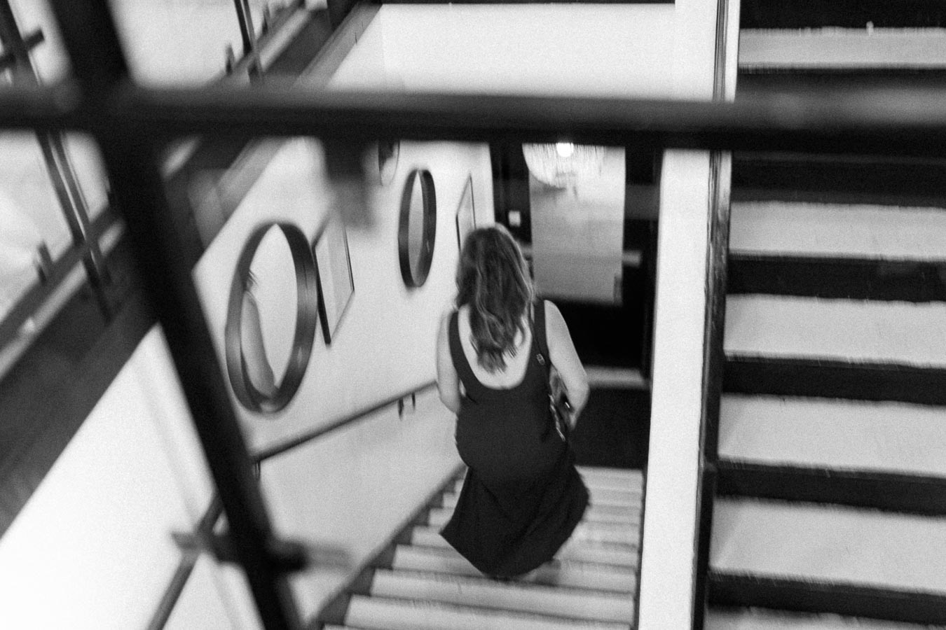 Black and white image of a woman in a dark dress descending a staircase, surrounded by modern decor and framed mirrors on the walls, capturing a candid moment in an elegant interior setting.