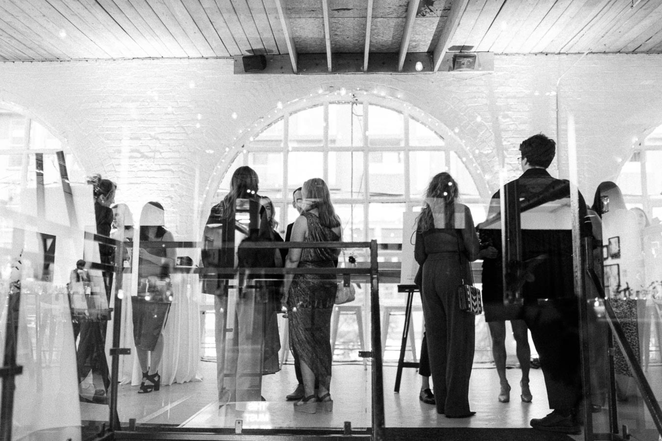 Black and white photo of a group of people socializing at an art gallery, with arched windows and modern decor creating a stylish, urban atmosphere.