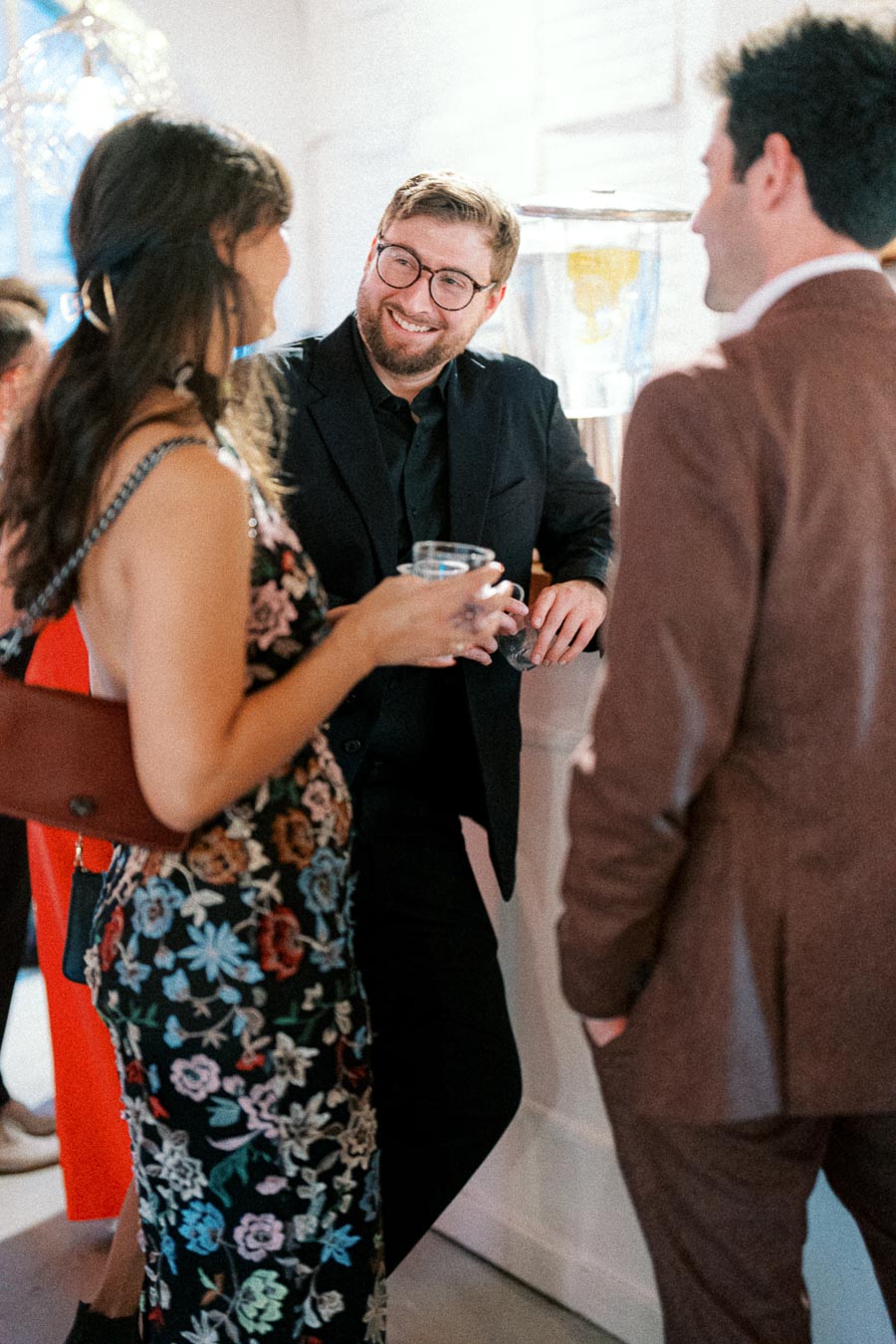 Group of three people engaging in conversation at a social event, with one man in glasses smiling and leaning against a counter while holding a drink. The woman is in a floral dress, and the other man wears a brown suit. Bright, well-lit indoor setting.