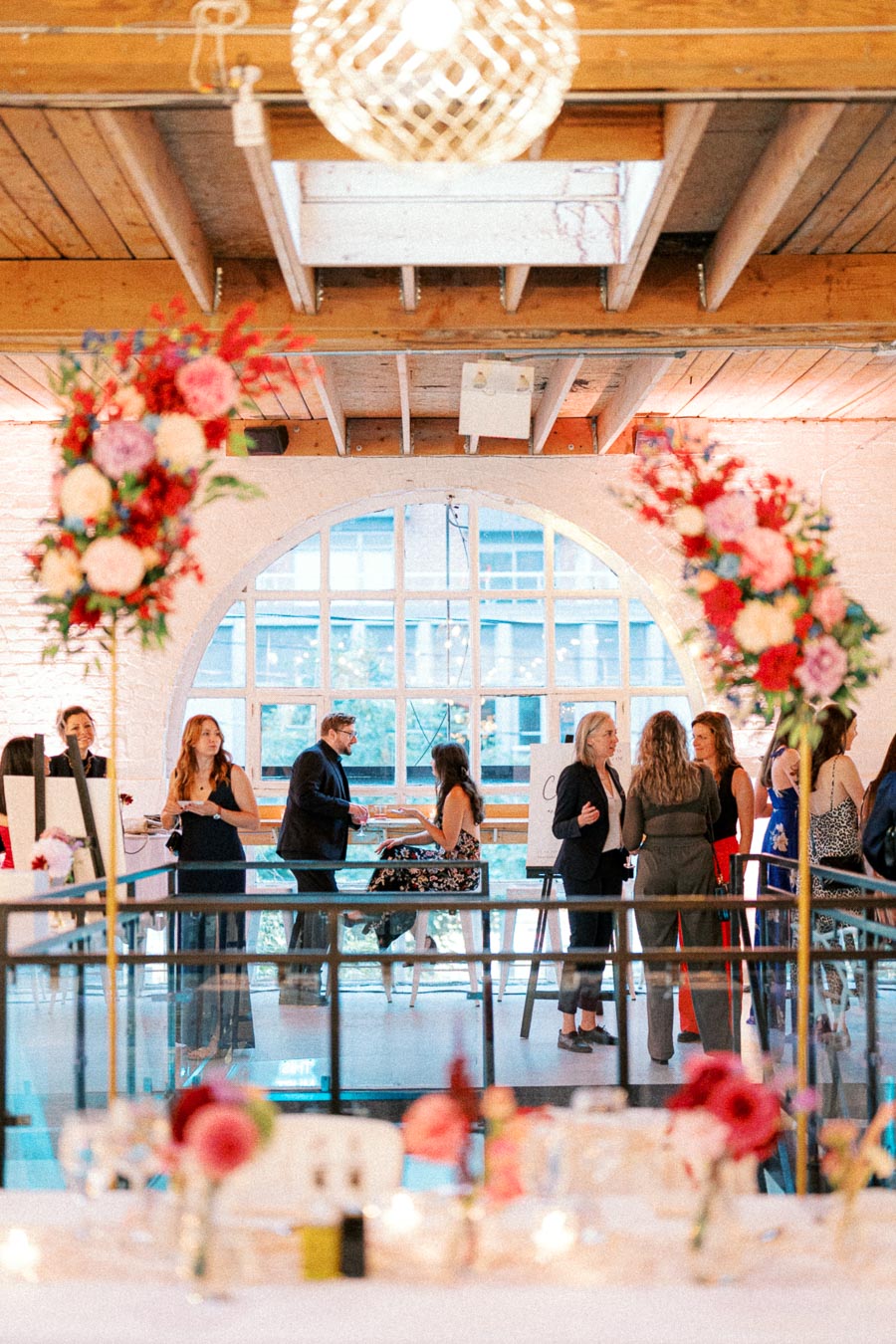 A vibrant indoor social event with elegantly dressed people mingling under a rustic wooden ceiling. Large floral arrangements frame the scene, and natural light pours through a large arched window, creating a warm and inviting atmosphere.