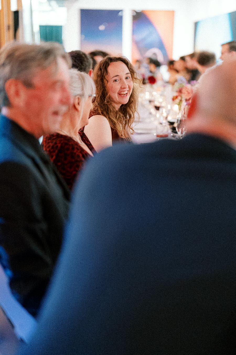 A joyful dinner gathering with a woman smiling brightly among a group of elegantly dressed guests seated at a long table, adorned with wine glasses and candles, in a warmly lit room.