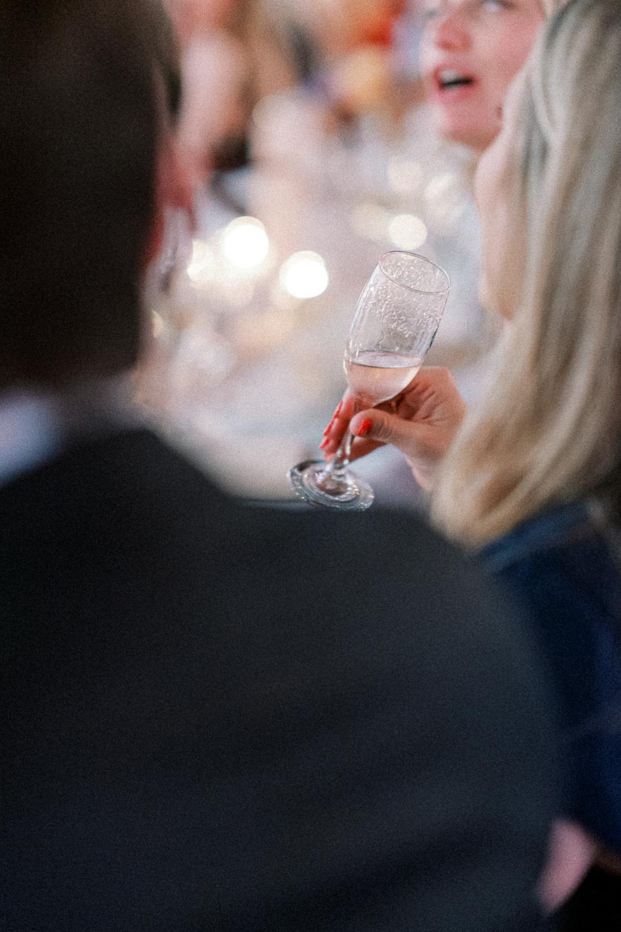 Elegant woman holding a champagne flute at a lively social event with softly lit background.