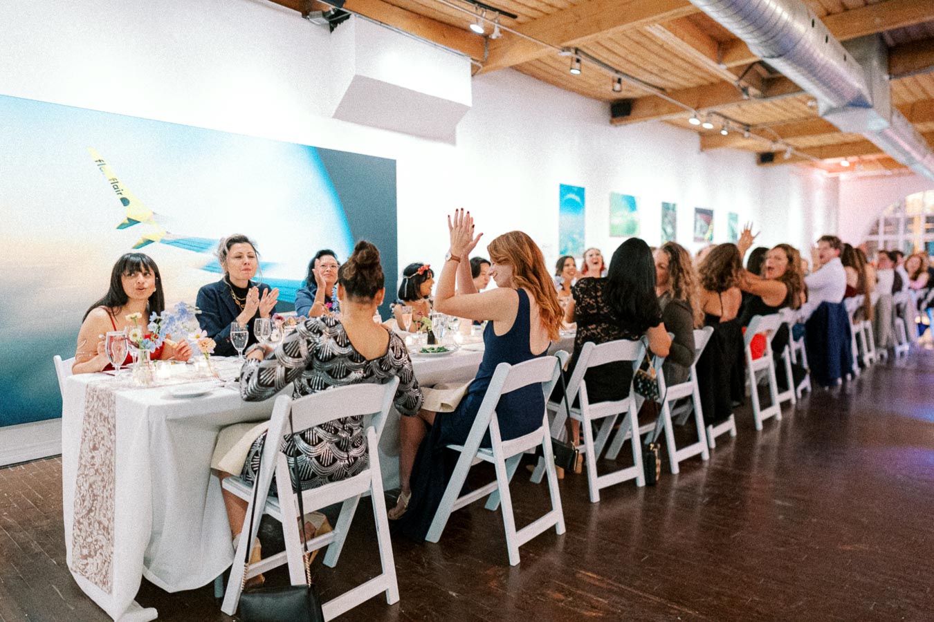 A large group of people sitting around a long table in a modern event space, engaged in conversation and laughter. The ambiance is lively with art on the walls and a wooden ceiling adding warmth to the venue.