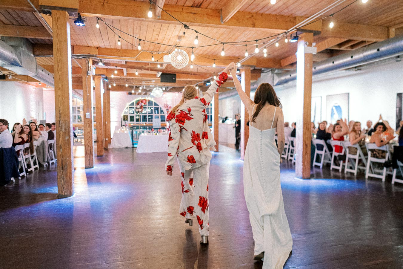 Celebratory dance in a rustic wedding reception venue with string lights, featuring someone in a floral suit and another in a flowing white dress.